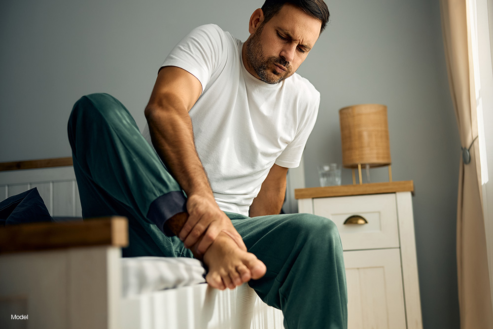 A man in green sweats sitting on a bed holding his foot in pain